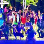 Grupo en fiesta nocturna junto a piscina con disfraces, DJ y bailarinas transmitiendo vibra festiva.