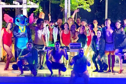 Grupo en fiesta nocturna junto a piscina con disfraces, DJ y bailarinas transmitiendo vibra festiva.