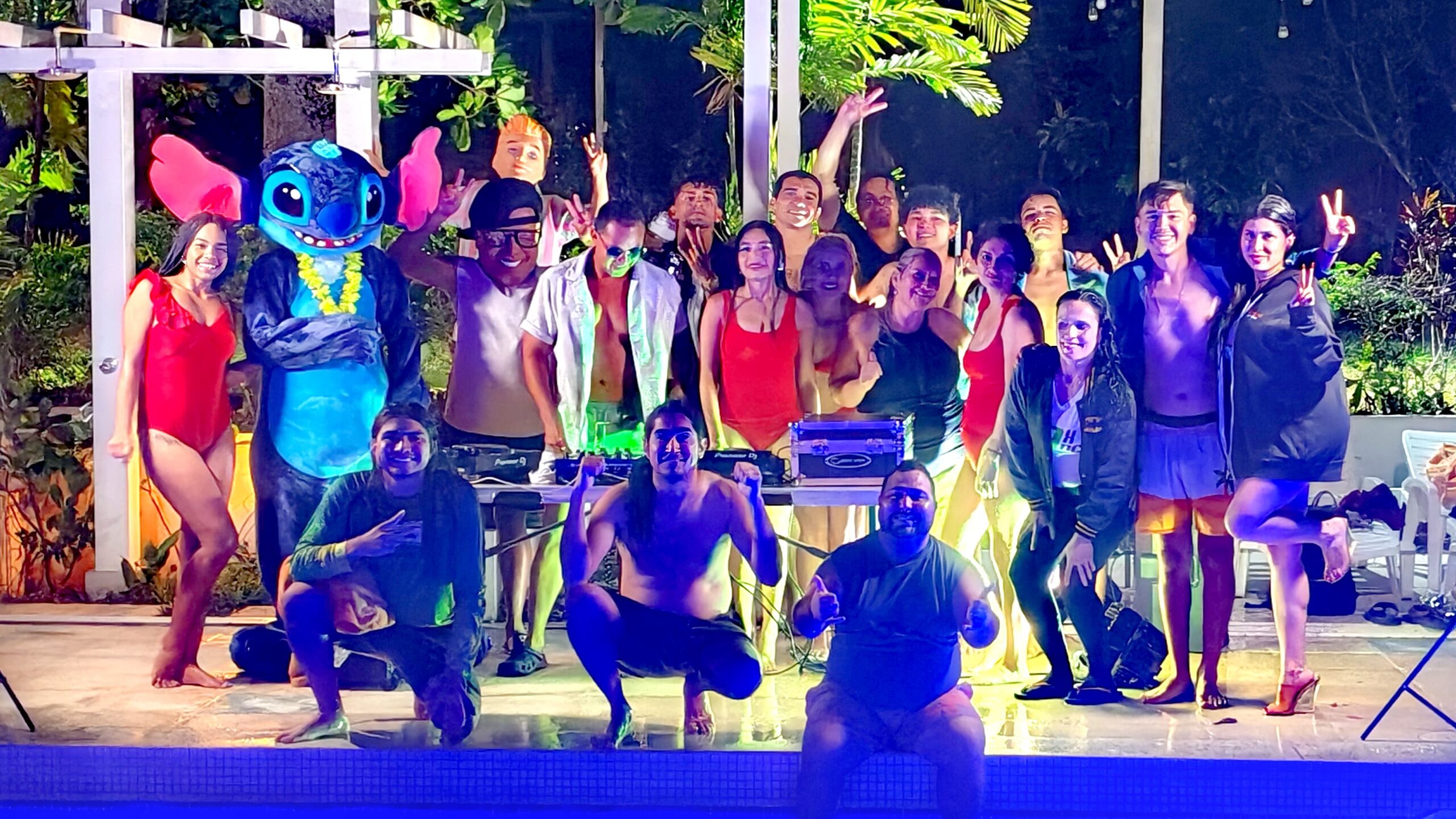 Grupo en fiesta nocturna junto a piscina con disfraces, DJ y bailarinas transmitiendo vibra festiva.
