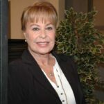 Retrato frontal de la actriz Gema Cuervo sonriendo, vistiendo un saco negro y blusa blanca con un collar dorado.