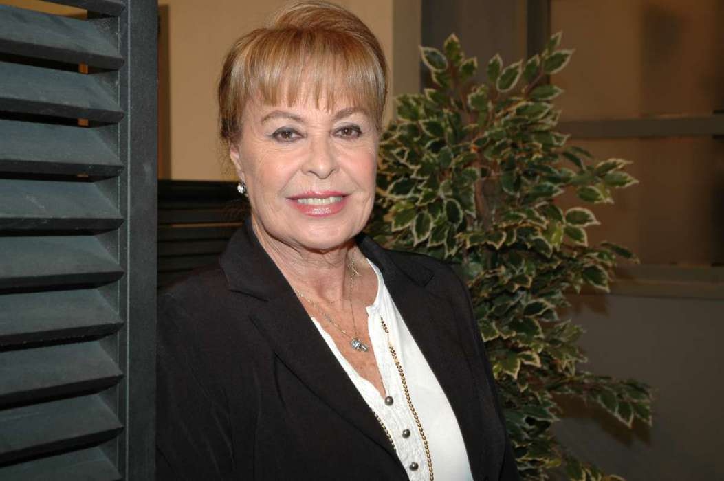 Retrato frontal de la actriz Gema Cuervo sonriendo, vistiendo un saco negro y blusa blanca con un collar dorado.